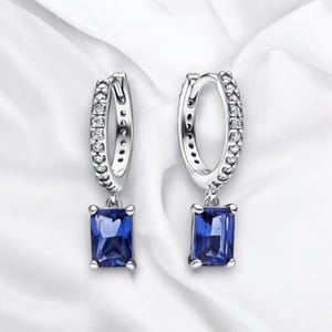 Op de afbeelding: Zilveren oorringen met een hangende blauwe edelsteen. De ring is versierd met kleine, heldere stenen.