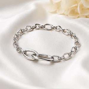 Può includere: Un bracciale a catena color argento caratterizzato da una serie di maglie ovali uniformi collegate a un meccanismo di chiusura ovale più grande e allungato. Il bracciale ha una finitura metallica lucida e riflettente.