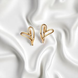 May include: A pair of gold-colored heart-shaped earrings. The earrings are made of a thin, curved metal that forms the shape of a heart. The earrings are displayed on a white, silky background.