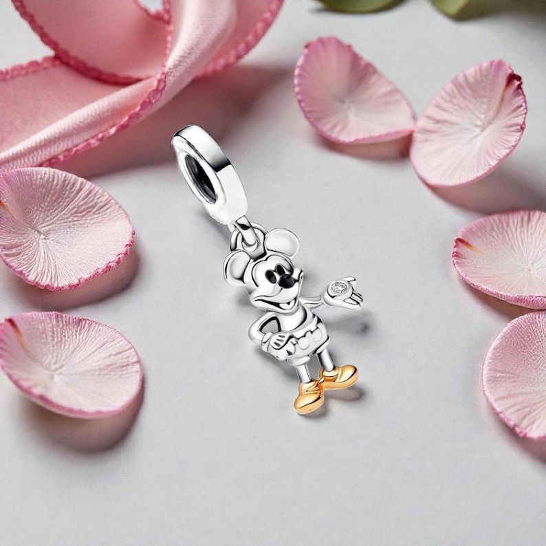 May include: A silver Mickey Mouse charm with gold-colored shoes and a small diamond accent. The charm is surrounded by pink flower petals and a pink ribbon. The charm is a pendant.