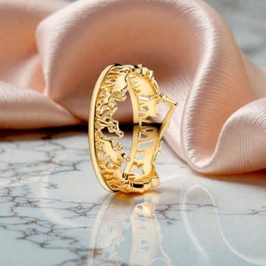 Può includere: Un anello dorato con un design traforato ornato. L'anello presenta ritagli dettagliati che creano un motivo decorativo. È presentato su uno sfondo di superficie marmorea e tessuto color pesca.