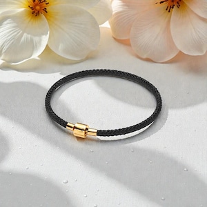 Puede incluir: Una pulsera circular de cordón negro trenzado con un cierre cilíndrico de metal en tono dorado pulido. La pulsera está colocada sobre una superficie blanca con sombras claras y rodeada de pétalos de flores blancas en las esquinas superiores.