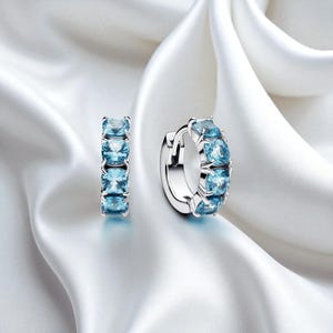 Op de afbeelding: Een paar zilveren oorringen, elk versierd met vijf vierkante, lichtblauwe edelstenen. De oorbellen worden gepresenteerd op een witte, zijdeachtige stof, waardoor de glans van de sieraden wordt benadrukt.