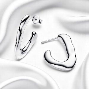 May include: A pair of silver-coloured hoop earrings. The earrings have an organic shape with a smooth, reflective surface. The earrings are displayed on a white, silky background.