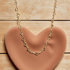 May include: A gold-colored necklace with heart-shaped links rests on a peach-colored heart-shaped cushion. The necklace is draped across the cushion, which is set against a light brown wooden surface.