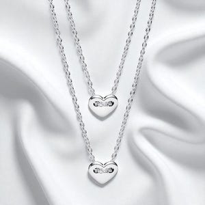 May include: Two silver necklaces with heart-shaped pendants. Each heart pendant has a small, decorative element in the center. The necklaces are displayed on a white, silky fabric, creating a soft, elegant look. The chains are delicate and the pendants are polished.