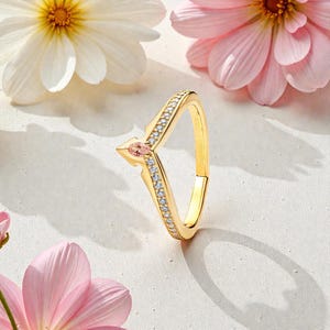 Pode incluir: Um anel dourado com um design único em forma de flecha. O anel apresenta uma pedra preciosa rosa central e é embelezado com pequenas pedras brancas brilhantes ao longo das bordas. O anel está sobre um fundo de flores rosa e brancas.