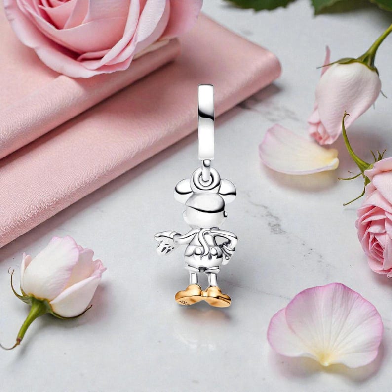 May include: A silver charm in the shape of a cartoon character with gold-colored shoes. The charm is suspended from a silver loop. Pink roses and petals are in the background.