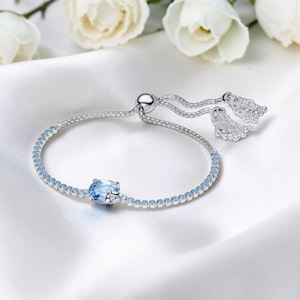May include: A silver bracelet with a delicate design. The bracelet features a central, light blue oval gemstone and is adorned with smaller, matching blue stones. The adjustable closure has two decorative, dangling elements.