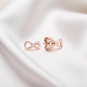 May include: Rose gold infinity stud earrings with small, clear gemstones. The earrings are displayed on a white, silky surface. The infinity symbol represents eternity and is a popular jewellery design.