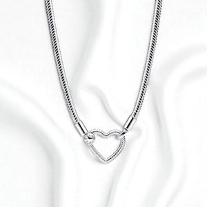 Op de afbeelding: Een zilveren ketting met een hartvormige hanger. De ketting heeft een getextureerd ontwerp en de hartvormige sluiting bevat een kleine, heldere steen. De achtergrond is een witte, zijdeachtige stof.