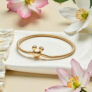 Op de afbeelding: Een goudkleurige armband met een Mickey Mouse-vormige sluiting. De armband is gemaakt van een flexibel, opgerold metaal en wordt gepresenteerd op een wit oppervlak, omringd door roze en witte bloemen.