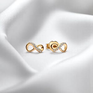 Op de afbeelding: Een paar goudkleurige oorstekers met een oneindigheidssymbool. Elke oorbel is versierd met kleine, heldere edelstenen. De oorbellen worden gepresenteerd op een witte, zijdeachtige achtergrond.