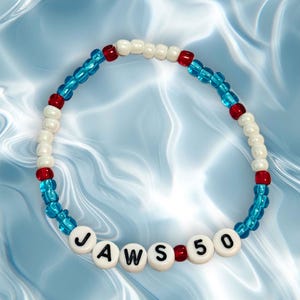 May include: A beaded bracelet with the text "JAWS 50". The bracelet features alternating sections of white, red, and blue beads. The beads are strung together to form a circular shape, suitable for wrist wear.