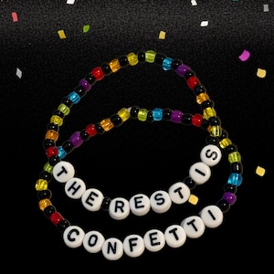 Op de afbeelding: Twee armbanden met kralen met de tekst "THE REST IS CONFETTI". De armbanden zijn gemaakt van kleurrijke kralen in regenboogtinten, met zwarte kralen die de kleuren scheiden. De achtergrond is zwart met confetti.