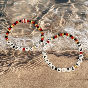 May include: Two beaded bracelets with white letter beads spelling "YOHO" and "PIRATES LIFE FOR ME". The bracelets have red, black, and gold beads. One bracelet features a skull bead.