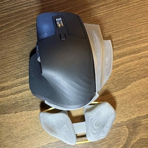 May include: A black Logitech computer mouse with a gray 3D-printed ergonomic support. The mouse has a scroll wheel and several buttons. The support is designed to cradle the user's wrist and fingers for comfort.