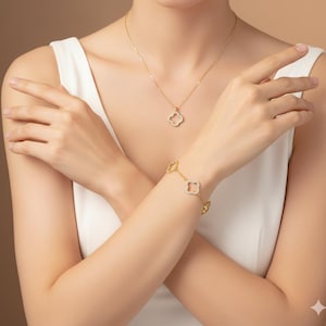 Op de afbeelding: Een gouden ketting en armband set met klavervormige hangers versierd met kleine, sprankelende stenen. De sieraden worden gedragen tegen een witte top, waardoor een contrast ontstaat dat de stukken benadrukt. De ketting hangt sierlijk, terwijl de armband om de pols wordt gedragen.
