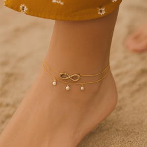 Puede incluir: Tobillera dorada con un símbolo de infinito y pequeños abalorios de perlas. La tobillera de cadena de doble capa se lleva en un tobillo, mostrando el delicado diseño de la joya. La tobillera es un accesorio de moda.