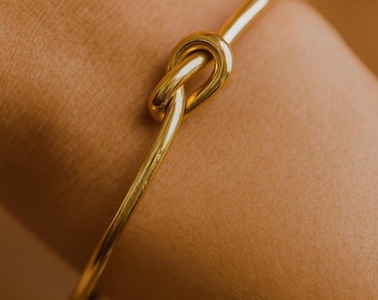 Peut inclure: Un bracelet jonc doré avec un motif de nœud. Le bracelet est en métal lisse et brillant et est porté au poignet. Le nœud est le point central, ajoutant un élément unique et élégant au bijou.