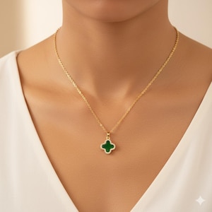 May include: A gold-colored necklace with a four-leaf clover pendant. The pendant is green with a border of small, clear stones. The necklace is worn against a white top.