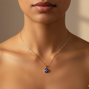 Puede incluir: Un collar dorado con un pequeño colgante redondo con un diseño de ojo azul. El colgante tiene un centro azul oscuro con un círculo blanco y un borde dorado. El collar se muestra en el cuello de una persona.
