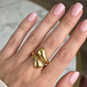 Può includere: Un anello color oro dal design aperto a bypass, caratterizzato da due estremità lisce a forma di goccia che si curvano attorno al dito. Il metallo ha una finitura lucida e riflettente, creando un aspetto elegante e continuo.