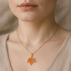 Puede incluir: Un collar dorado con un colgante de hoja de arce naranja. El colgante tiene un borde dorado. El collar se lleva alrededor del cuello.