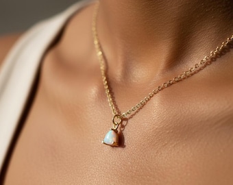 May include: A gold necklace with a small, triangular pendant containing an iridescent opal. The pendant hangs from a fine gold chain, offering a minimalist and elegant look. The necklace is displayed against a skin tone background.