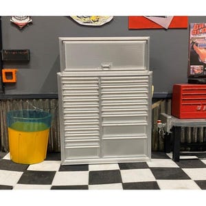 Puede incluir: Un cofre de herramientas plateado con múltiples cajones y un compartimento superior. El cofre está colocado contra una pared gris con un suelo a cuadros en un entorno de garaje. Una taza amarilla y azul está a la izquierda.