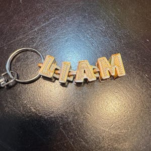 May include: A gold-colored keychain spelling out the name "LIAM". The letters are in a block style with a textured surface and are connected by thin metal bars. The keychain has a silver-colored ring.