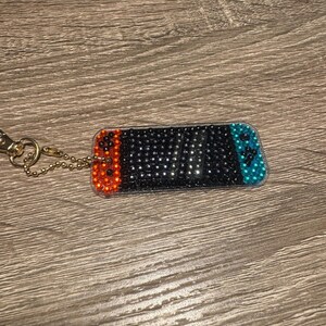 Könnte beinhalten: Ein schwarzer und silberner Nintendo Switch Schlüsselanhänger mit orangefarbenen und blauen Strasssteinen. Der Schlüsselanhänger ist an einer goldenen Kette befestigt.