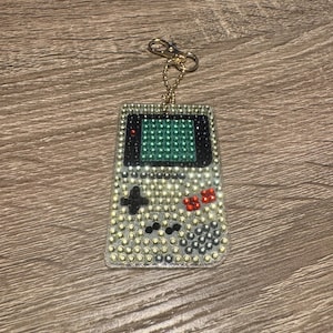 Könnte beinhalten: Ein Schlüsselanhänger in Form einer alten tragbaren Videospielkonsole aus klarem Kunststoff mit einem grünen Bildschirm und schwarzen Knöpfen. Der Schlüsselanhänger ist mit kleinen, bunten Strasssteinen besetzt.