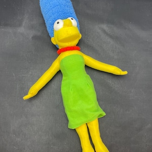 May include: A plush toy of Marge Simpson from The Simpsons. She is wearing a green dress and red shoes. Her hair is blue and her skin is yellow.