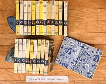 May include: A collection of Nancy Drew mystery books by Carolyn Keene with yellow spines and Grosset & Dunlap branding. One open book displays blue and white patterned endpapers featuring various illustrations. A white label reads YELLOW MATTE BOOKS WITH BLUE ENDPAPERS.