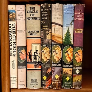 May include: A collection of vintage Carolyn Keene mystery books, including titles like "The Secret at Lone Tree Cottage" and "The Winking Ruby Mystery." The spines display colorful illustrations and text, with the series name "The Dana Girls Mystery Stories" visible on some.