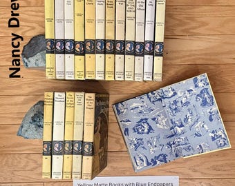May include: A collection of Nancy Drew mystery books by Carolyn Keene, featuring yellow spines with black text and blue circular illustrations. One book is open to reveal blue and white patterned endpapers. Two grey rock bookends are included.