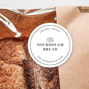 May include: A white round sticker with black text that reads "Freshly Baked Sourdough Bread The Sourdough Girl" on a brown paper bag with a loaf of sourdough bread inside.