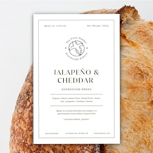 Könnte beinhalten: Ein Sauerteigbrot mit einem Etikett mit der Aufschrift "Jalapeño & Cheddar". Das Etikett zeigt das Logo der Bäckerei, die Zutaten und ein Nettogewicht von 900g. Das Brot hat eine goldbraune Kruste.