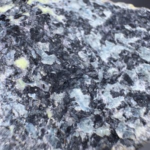 May include: Close-up of a rough, textured rock formation. The stone features a mix of dark gray, black, and pale blue-green colors. The surface has a crystalline appearance with areas of light reflection, suggesting a mineral specimen.