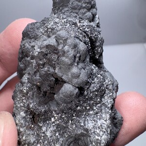 May include: A dark gray mineral specimen with a rough, textured surface. The rock has a crystalline structure with small, shiny, silver-colored inclusions throughout. The mineral is held in a hand.