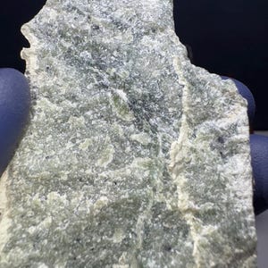 Puede incluir: Primer plano de una piedra toscamente labrada, de forma irregular. La piedra presenta un aspecto moteado con tonos verdes, grises y blancos. Pequeños puntos oscuros están dispersos por la superficie. La textura parece áspera y desigual.
