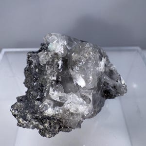 May include: A raw, uncut crystal cluster with a mix of clear and opaque sections. The crystal has a rough, textured surface with a dark gray matrix. The overall shape is irregular, showcasing natural geological formations.