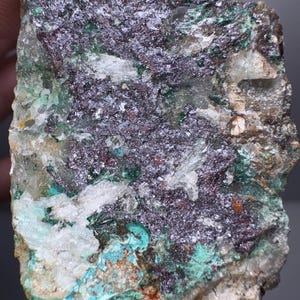 Könnte beinhalten: Nahaufnahme einer rohen Mineralprobe mit einer Mischung aus Farben. Der Stein weist Bereiche in tiefem Lila, Türkis und Grün auf, mit Flecken in Weiß und Hellbraun. Die Oberfläche hat ein raues, strukturiertes Aussehen.