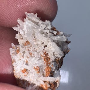 May include: A small cluster of white, needle-like crystals on a brown matrix. The crystals are clustered together in a dense, almost fluffy formation.