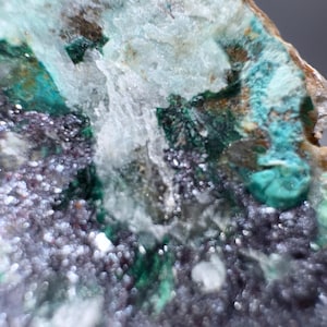 May include: Close-up of a mineral specimen with a mix of colors. The image shows a combination of teal, green, white, and dark purple hues. The mineral has a crystalline structure with a rough, textured surface.