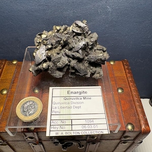 Könnte beinhalten: Ein graues Enargit-Mineralienstück aus der Mine Quiruvilca in Peru. Das Stück wird auf einer Holzkiste mit einem Etikett präsentiert, auf dem steht: "Enargit, Quiruvilca Mine, Quiruvilca Division, La Libertad Dept, Peru, Acc. No. 1094, Hey No. 06.03.01, W. A. BOLTON COLLECTION".