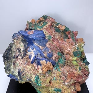 May include: A colorful mineral specimen with blue, green, and red crystals. The specimen is on a black display stand.