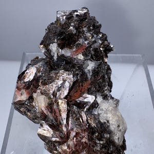 May include: A close-up of a mineral specimen with a dark, layered structure. The mineral has a mix of dark brown, silver, and reddish-brown hues, with crystalline formations and patches of white quartz.