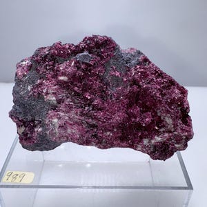 Puede incluir: Un espécimen mineral en bruto de color magenta intenso, intercalado con parches grises oscuros y blancos. La superficie rugosa y texturizada de la roca se exhibe sobre un soporte de acrílico transparente. Una pequeña etiqueta muestra el número "989".